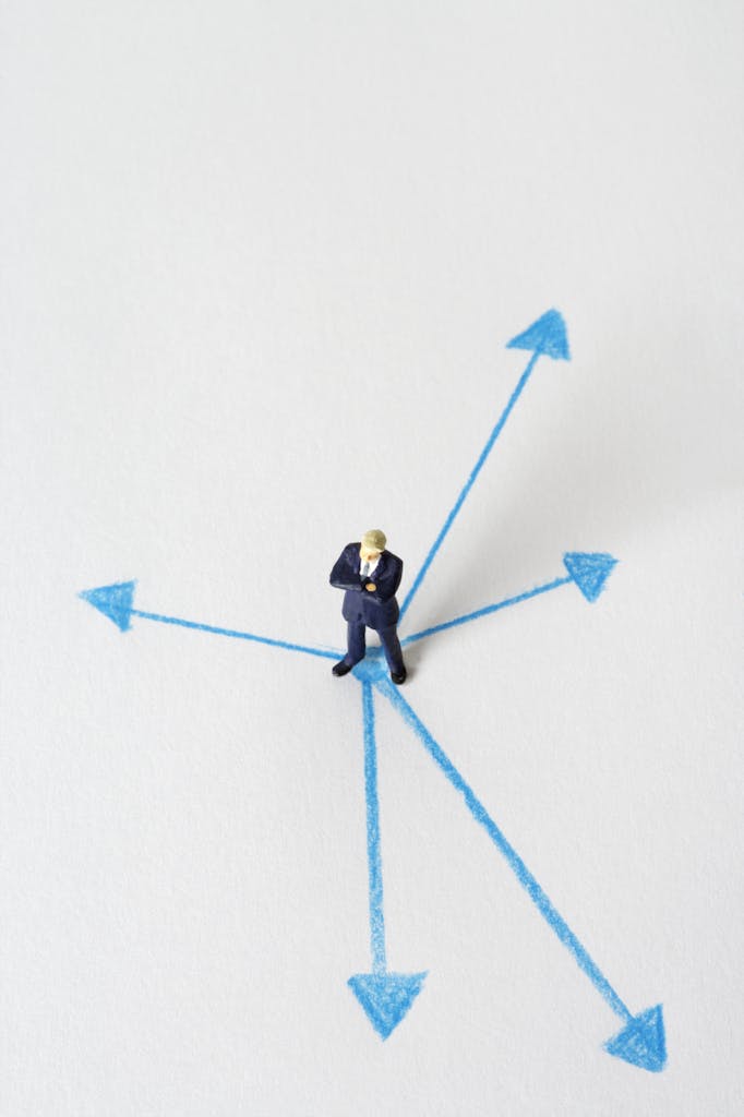 Businessman at Decision Crossroads | Symbolic Leadership & Strategy Photo. Miniature businessman standing at a crossroads of blue arrows on a white background, symbolizing decision-making, career paths, or business strategy choices.