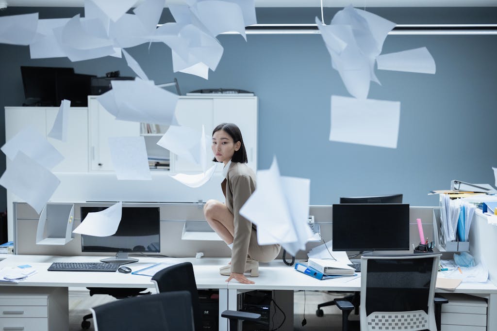 Excel Spreadsheet Chaos - Business Stress and Workplace Inefficiency. Stressed businesswoman sitting on desk surrounded by flying papers in chaotic office, representing Excel spreadsheet overwhelm and workplace inefficiency.